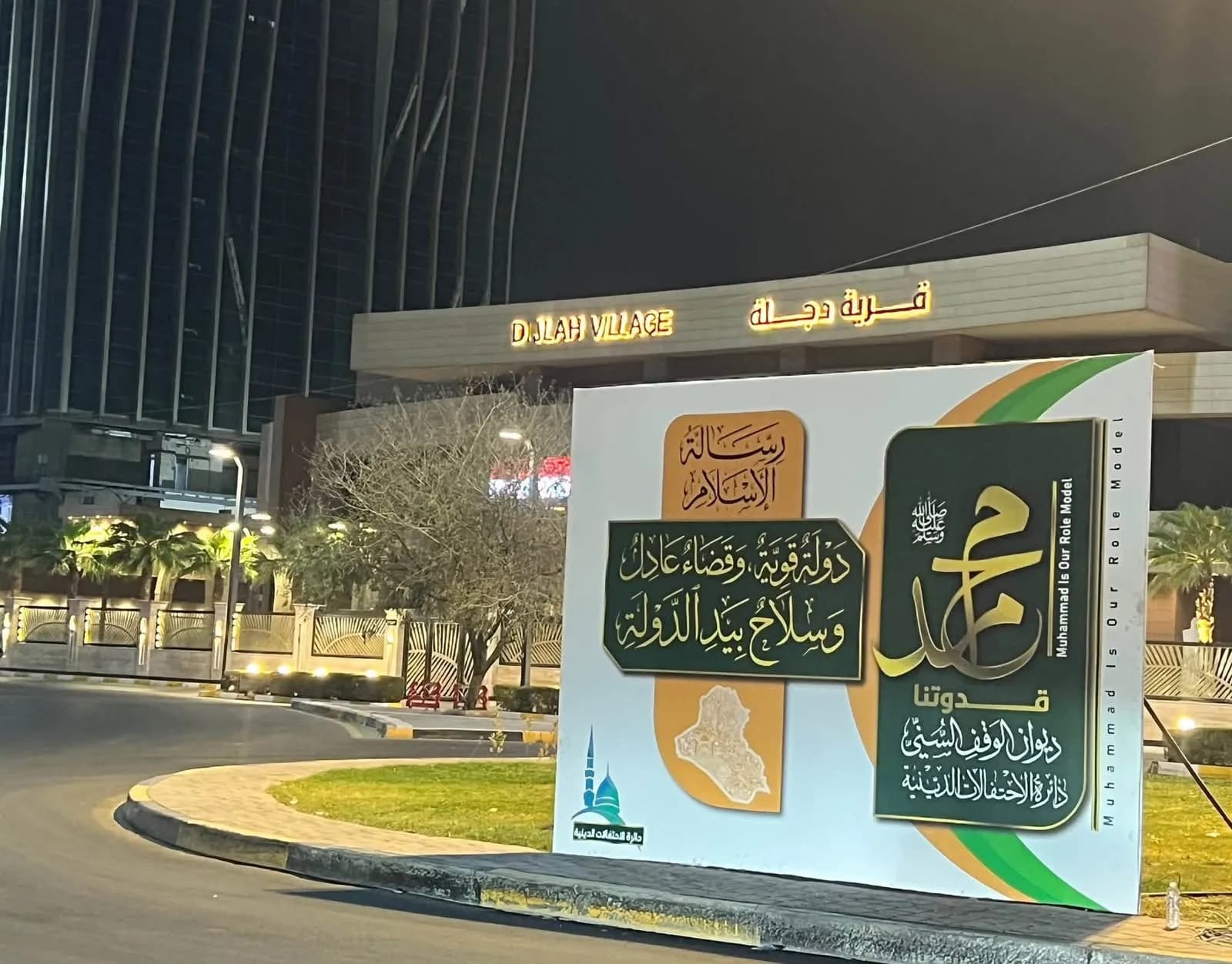 صورة لافتة ترويجية مضاءة ليلاً أمام مدخل قرية دجلة، مكتوب عليها "محمد قدوتنا" باللغتين العربية والإنجليزية.