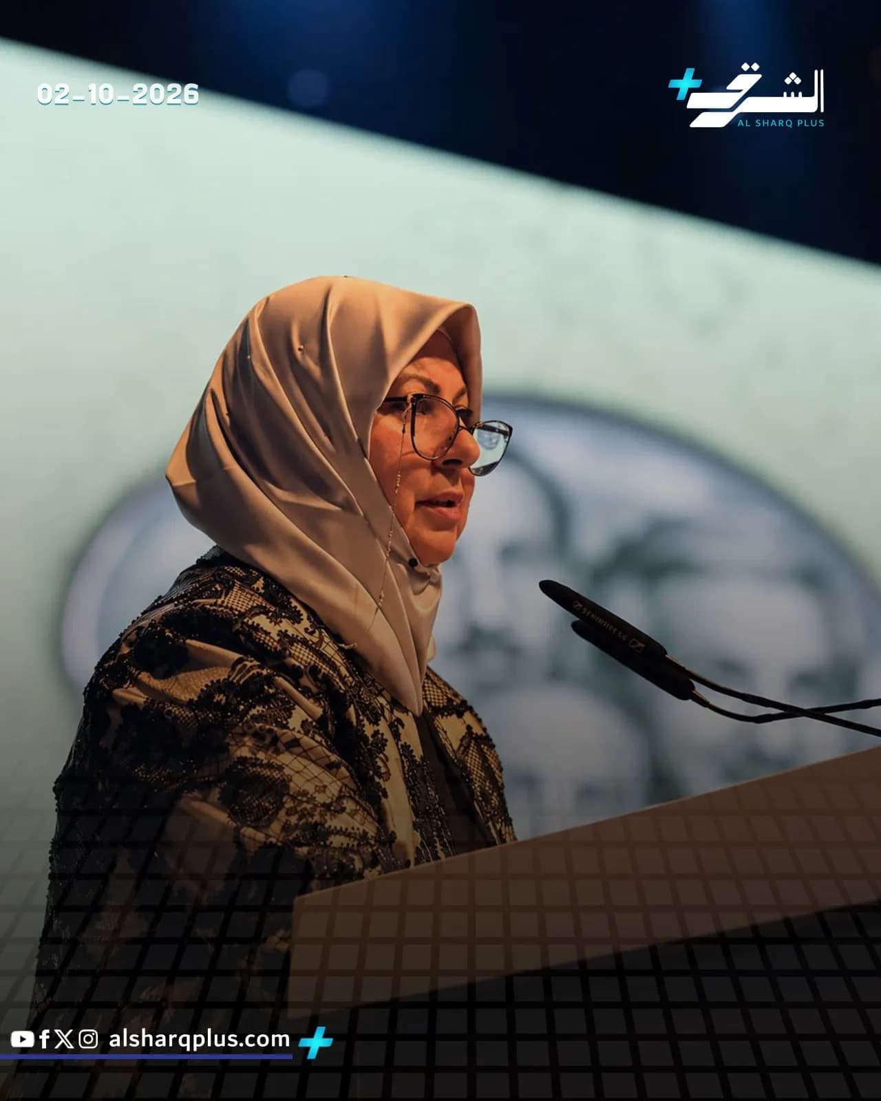 سيدة ترتدي حجابًا ونظارة تقف خلف منبر وتتحدث، وعلى يسار الصورة تاريخ "02-10-2026".