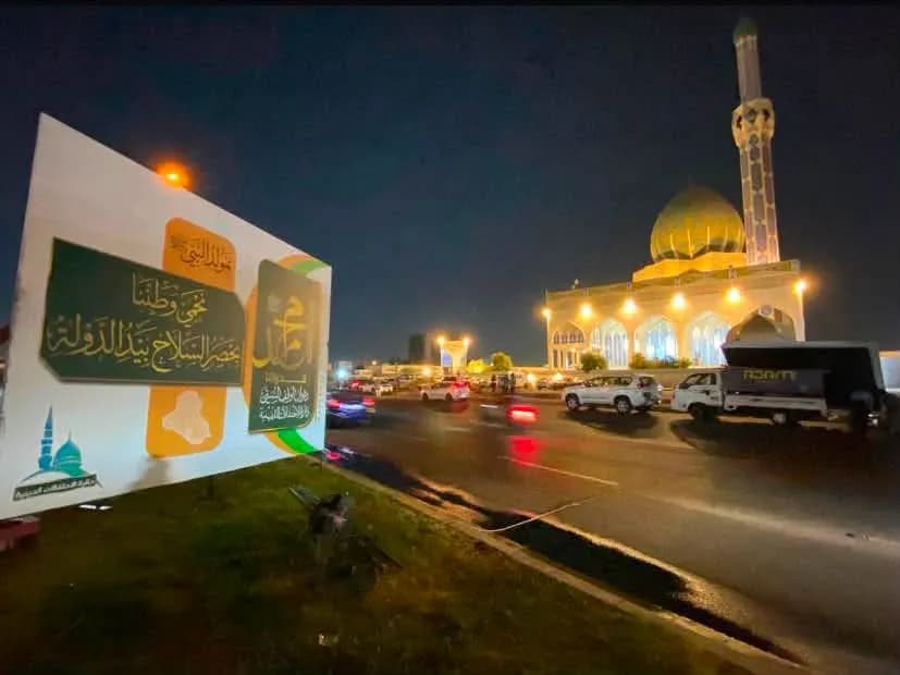 صورة ليلية لمسجد بقبة ذهبية ومئذنة عالية مضاءة، بجواره طريق تظهر عليه حركة مرور.