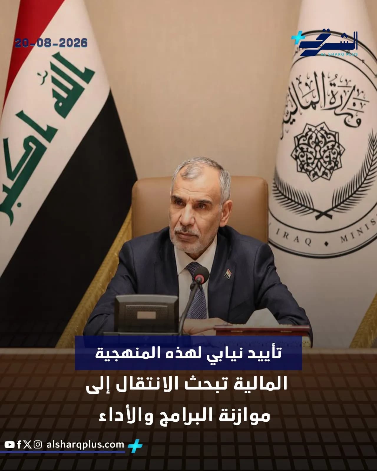 رجل يرتدي بدلة داكنة وربطة عنق زرقاء يجلس أمام علمين في مكتبه، أحدهما علم العراق.