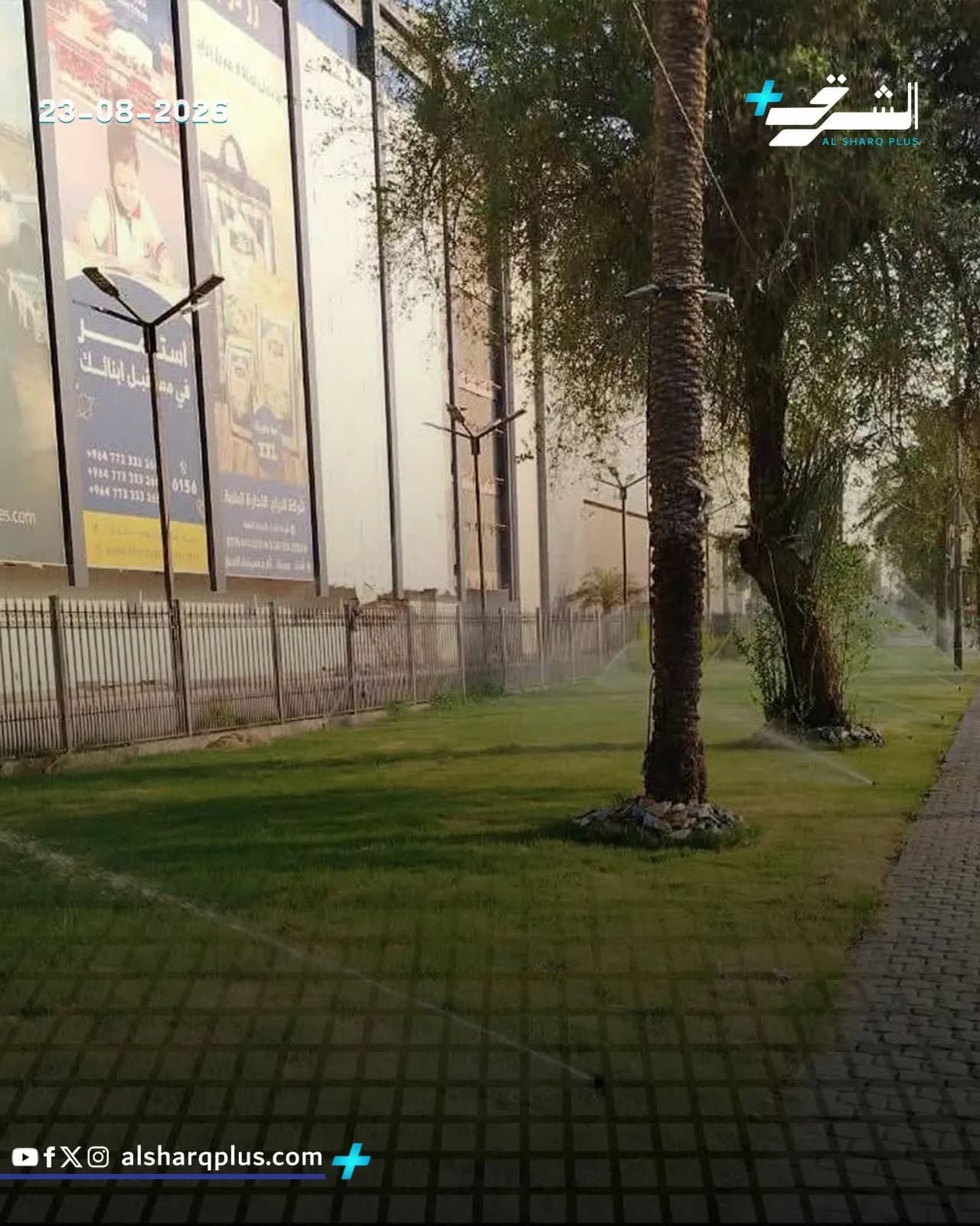 منظر جانبي لمبنى كبير يضم لوحات إعلانية، مع طريق مبلط وحشائش خضراء تُروى برشاشات مياه.