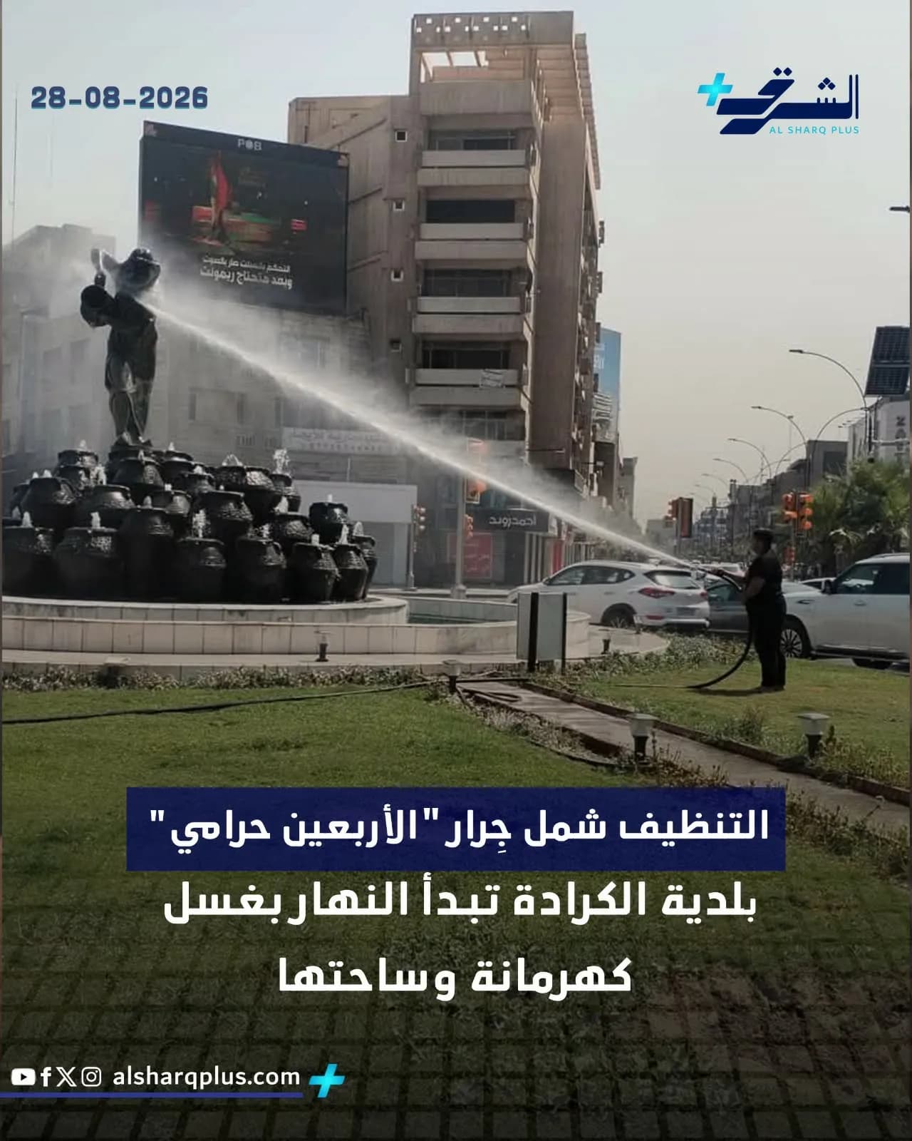 رجال البلدية يغسلون تمثالاً في ساحة الكرادة ببغداد، مع رشاشات مياه وسيارات على الطريق، ومبنى شاهق في الخلفية.