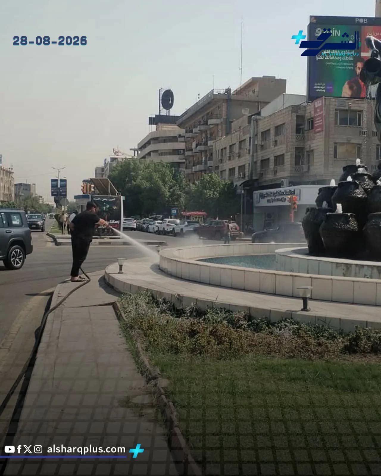 رجل يسقي بالخرطوم جانب طريق بجانب نافورة كبيرة في منطقة حضرية مليئة بالمباني.
