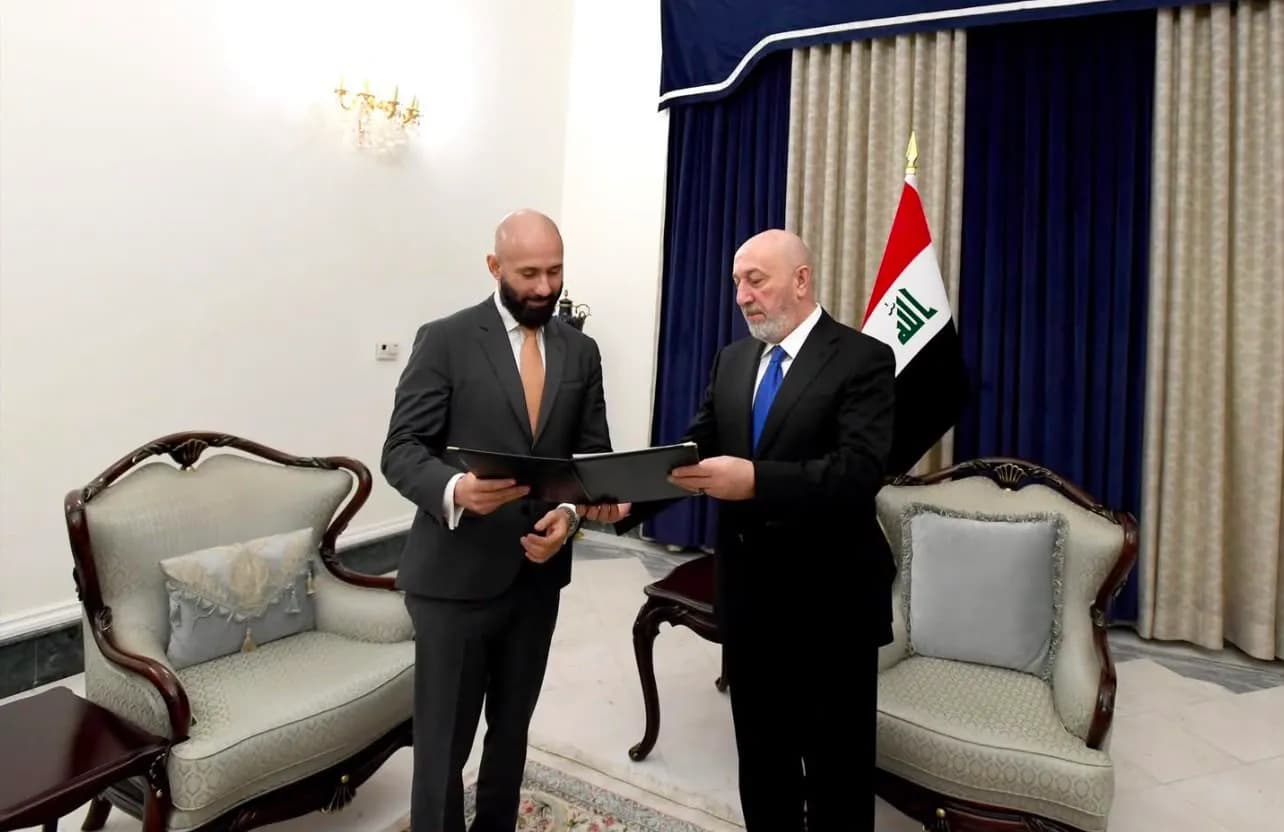 مسؤولان عراقيان يرتديان بدلتين رسميتين يتبادلان ملفات، ويقفان في غرفة مكتب أمام علم العراق.
