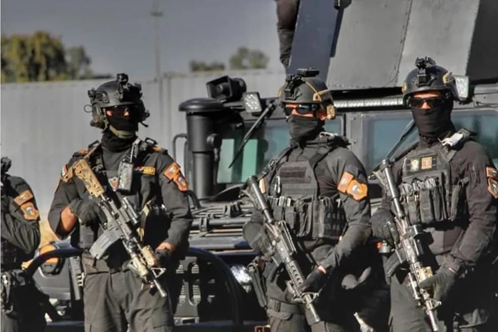 ثلاثة رجال يرتدون زيًا عسكريًا أسود وخوذات ونظارات شمسية ويحملون بنادق، يقفون أمام عربة مدرعة.