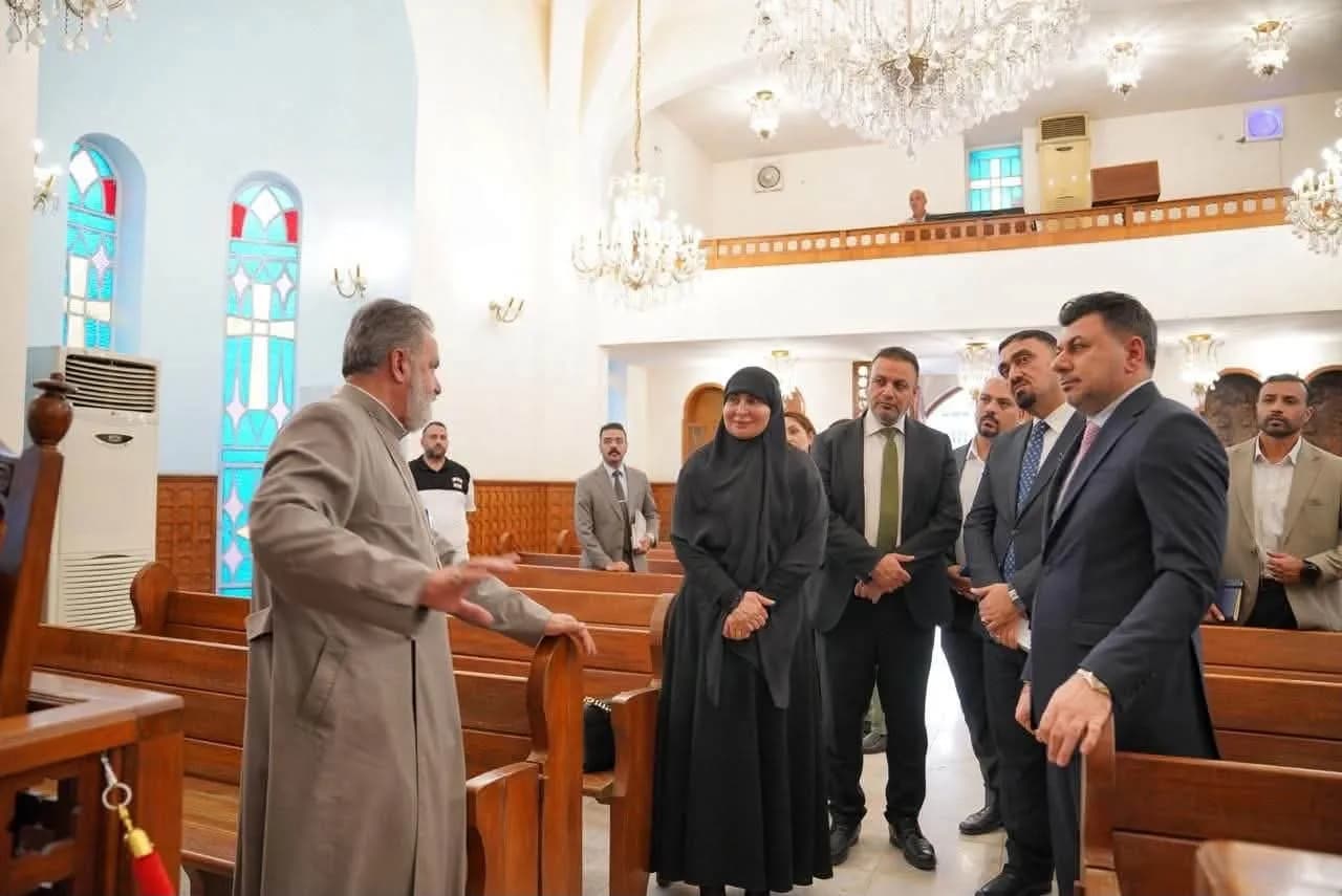 مجموعة من الرجال والنساء يرتدون ملابس رسمية داخل ما يبدو أنه كنيسة مزينة بنوافذ زجاجية ملونة وثريات.