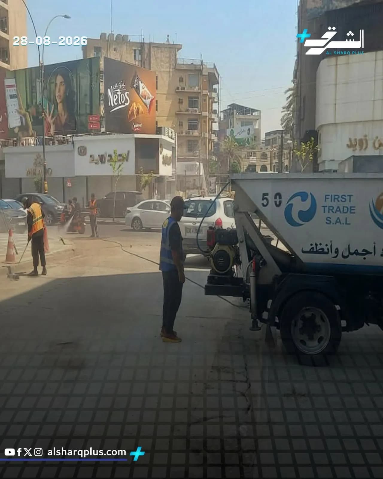عمال ينظفون شارعًا في المدينة بمرشات مياه، مع وجود لافتات إعلانية ومباني في الخلفية.